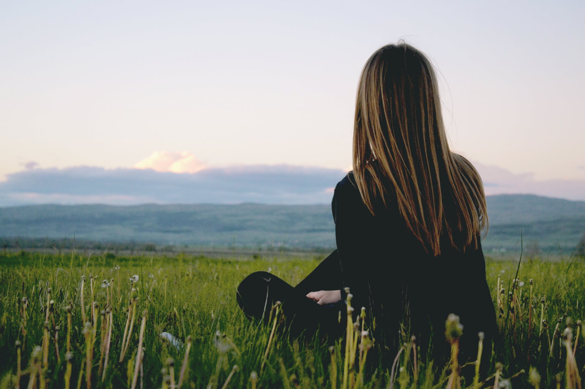 woman wearing black long sleeved shirt sitting on green grass field near mountain under cloudy sky